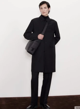 A person in a black overcoat and pants with a messenger bag, standing against a cream-colored wall and brown baseboard, studio shot.