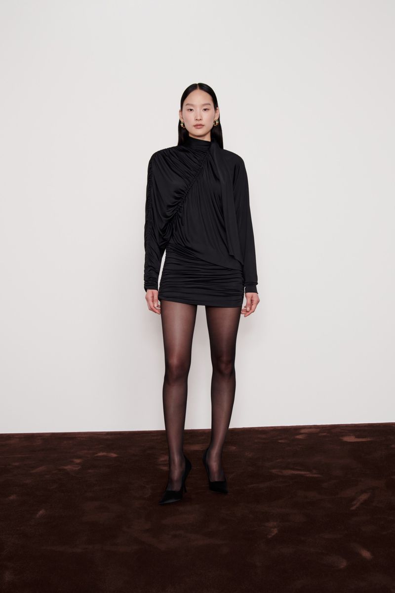 A full studio shot presents a slender woman in a black mini dress and tights, set against a clean white backdrop with a brown carpet.