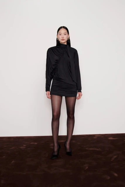 A full studio shot presents a slender woman in a black mini dress and tights, set against a clean white backdrop with a brown carpet.