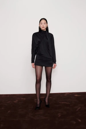 A full studio shot presents a slender woman in a black mini dress and tights, set against a clean white backdrop with a brown carpet.