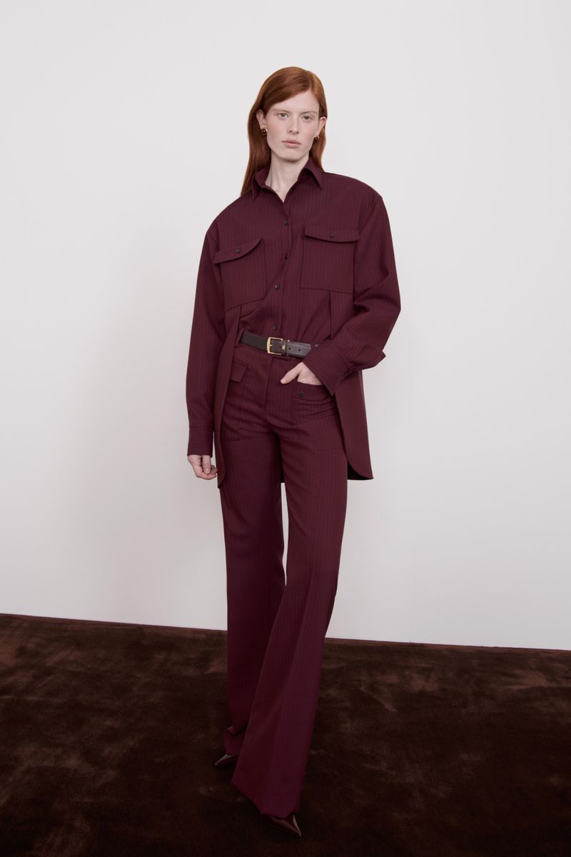 A red-haired woman is posing in a maroon-colored long-sleeved shirt and flared pants with a brown belt against a neutral background.