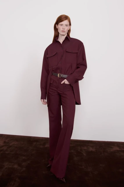 A red-haired woman is posing in a maroon-colored long-sleeved shirt and flared pants with a brown belt against a neutral background.