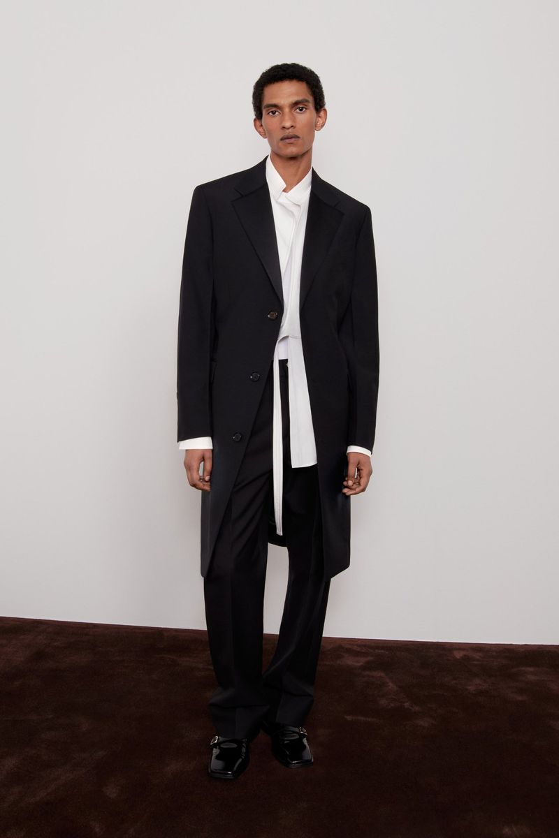 Elegant adult male fashion model in a black coat, white shirt, and black pants standing in a photo studio with a light background.