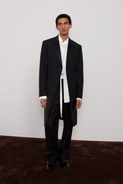 Elegant adult male fashion model in a black coat, white shirt, and black pants standing in a photo studio with a light background.