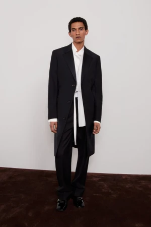 Elegant adult male fashion model in a black coat, white shirt, and black pants standing in a photo studio with a light background.