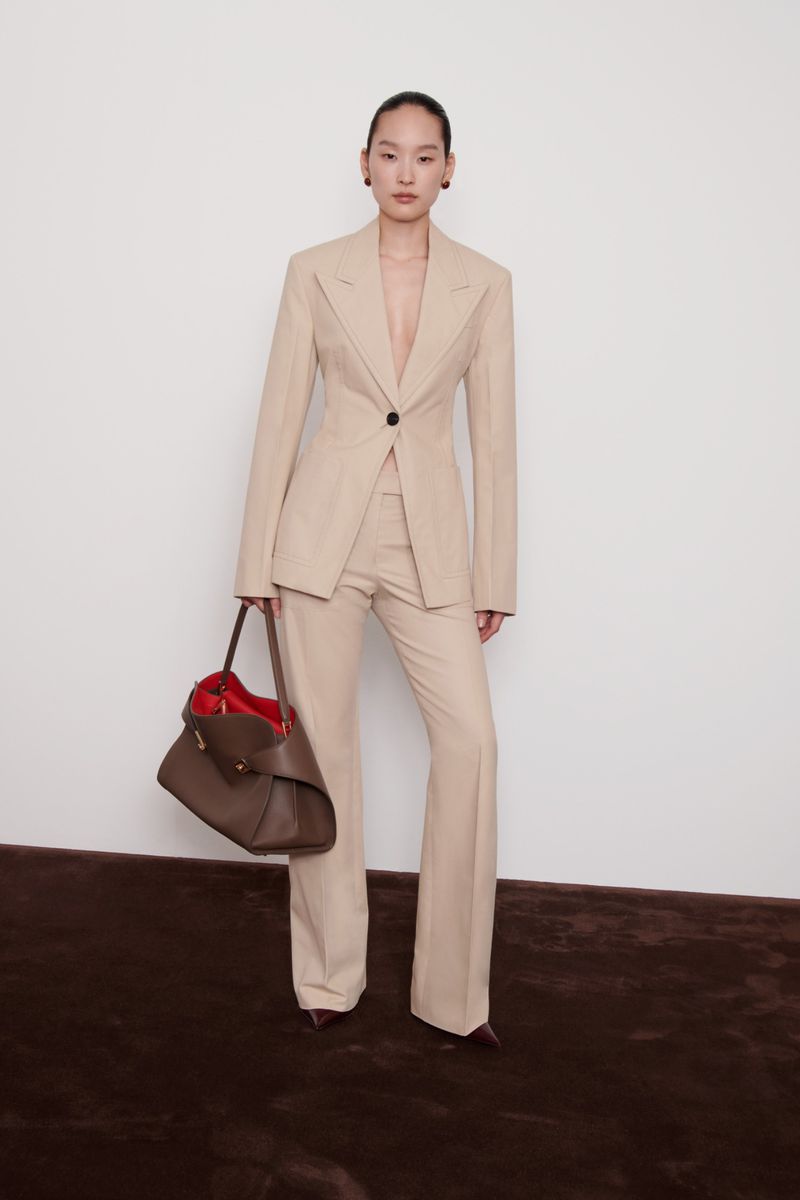 Elegant full-length shot of an Asian female model in a tan pantsuit holding a brown handbag against a white wall and dark brown floor.