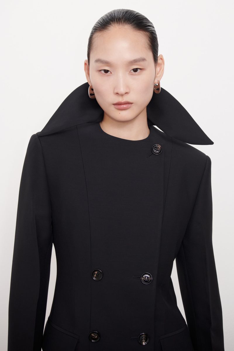 Headshot of a stylish Asian woman wearing a black double-breasted blazer with a large collar and earrings against a white backdrop.