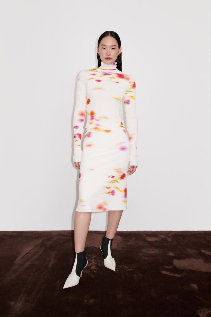 An Asian adult woman is posing in a long-sleeve white dress with colorful abstract patterns and white and black heeled boots on a brown surface.