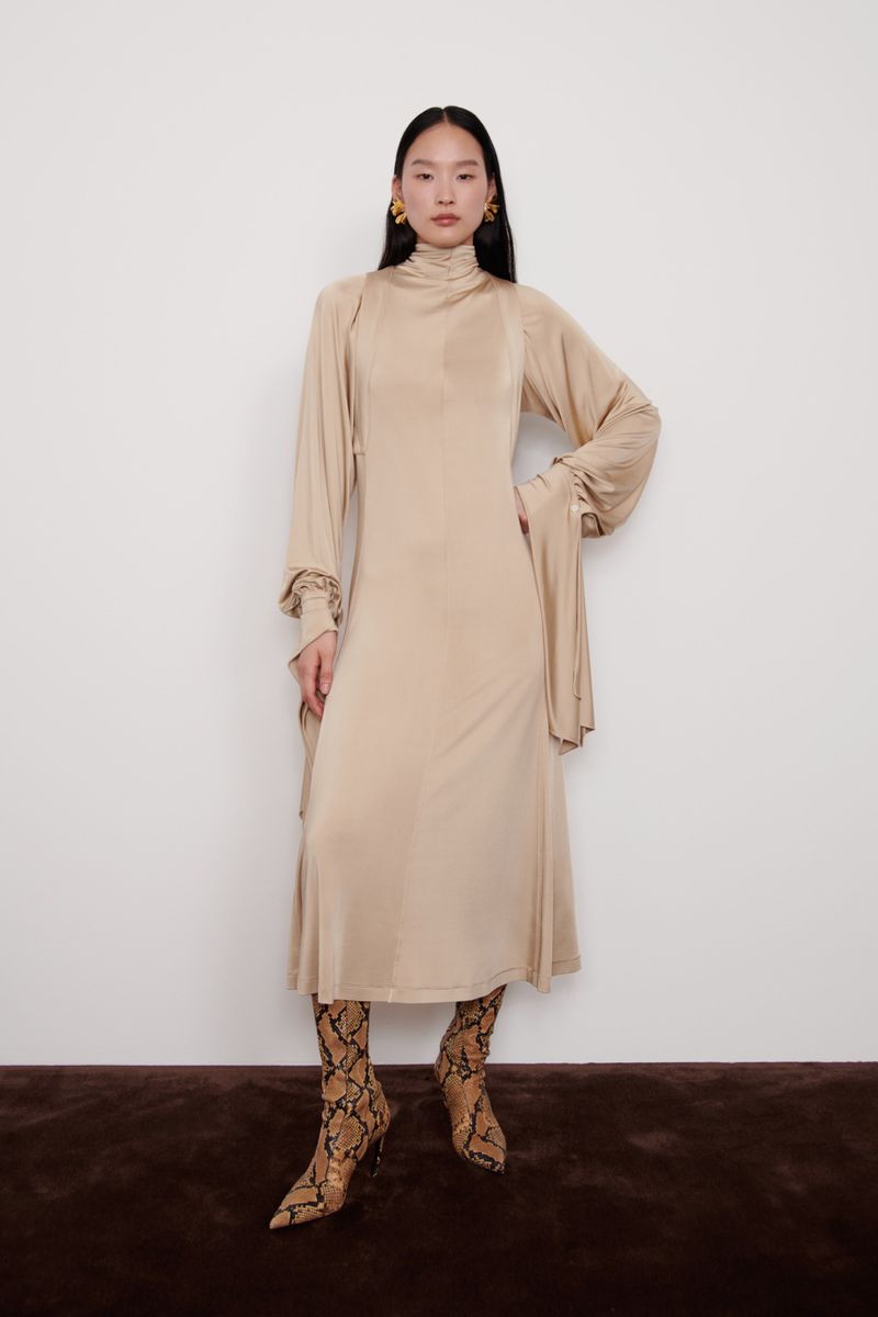 Asian woman in a beige dress and snake print boots posing against a white wall, fashion editorial shot. Long sleeve dress with high neck.