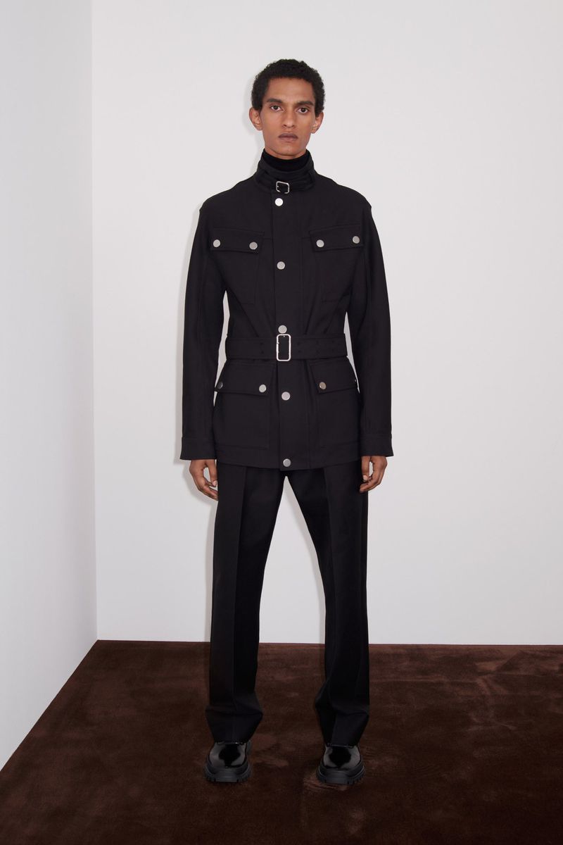 Fashion shot of a man in a black belted safari jacket and tailored pants, posing against a plain white wall with a brown floor.