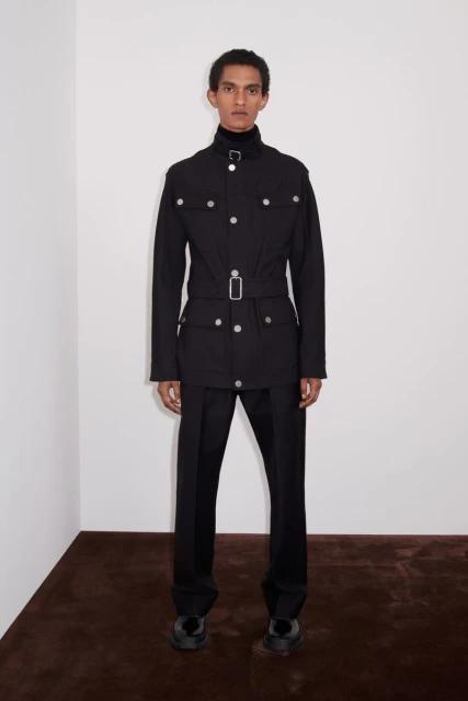 Fashion shot of a man in a black belted safari jacket and tailored pants, posing against a plain white wall with a brown floor.