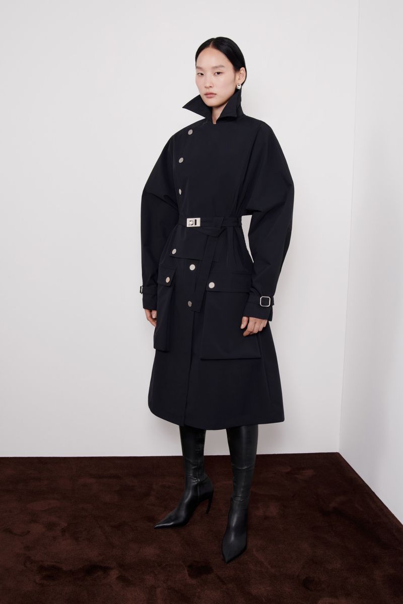 A woman models a black trench coat and leather boots against a white wall and brown floor, showcasing a blend of modern and classic style.