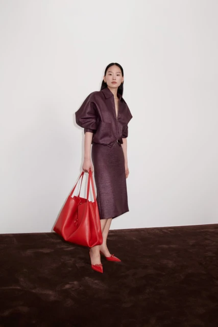 A woman poses in a brown skirt and top holding a large red tote bag in a studio with a white wall and brown floor.