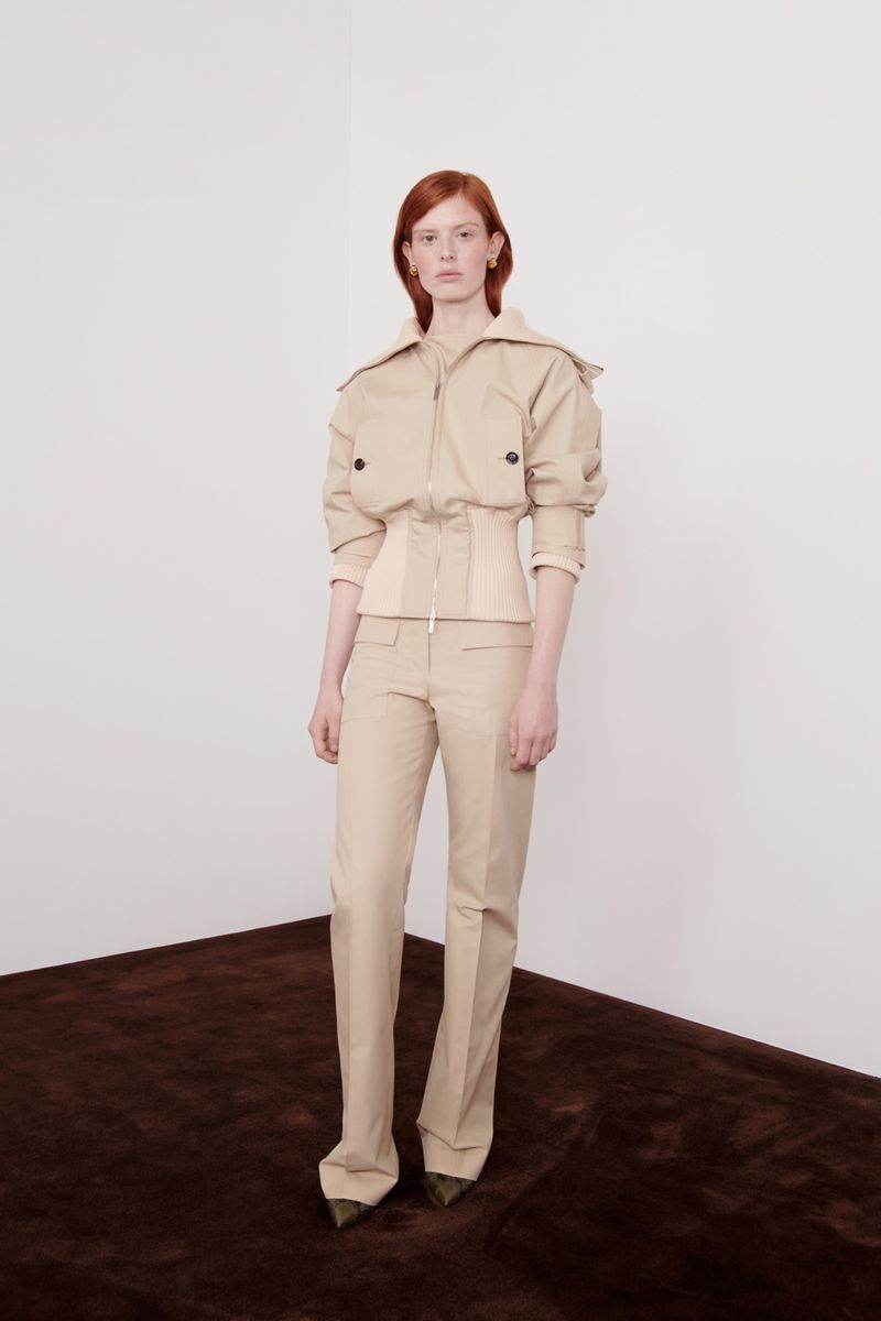 A woman with red hair poses in a beige jacket and pants in a studio setting with a dark brown carpet and a white wall.