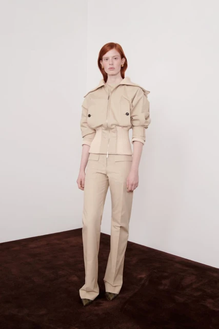 A woman with red hair poses in a beige jacket and pants in a studio setting with a dark brown carpet and a white wall.
