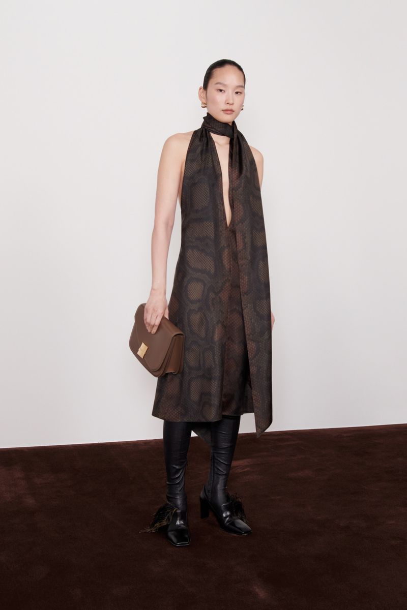 Fashion shot of an Asian woman wearing a snakeskin-pattern dress, black boots, and a brown purse in front of a white wall.