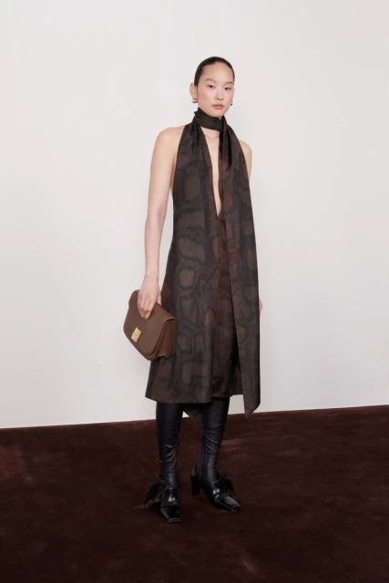 Fashion shot of an Asian woman wearing a snakeskin-pattern dress, black boots, and a brown purse in front of a white wall.