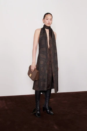 Fashion shot of an Asian woman wearing a snakeskin-pattern dress, black boots, and a brown purse in front of a white wall.