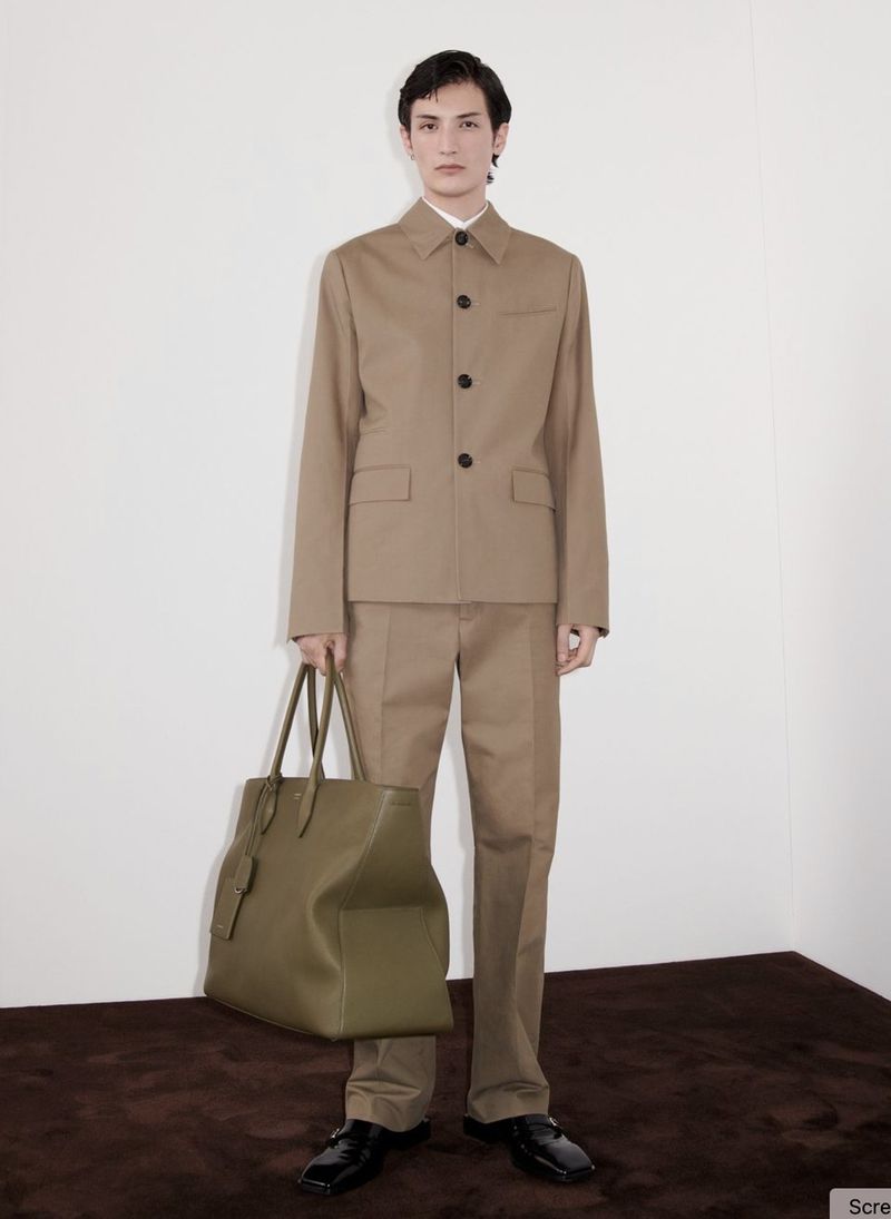 A stylish adult male model poses indoors wearing a tan suit, black shoes, and holding a large olive-green tote bag, in a fashion shoot.