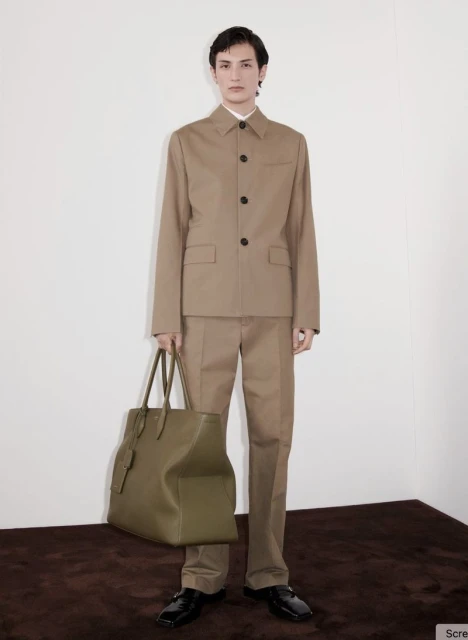 A stylish adult male model poses indoors wearing a tan suit, black shoes, and holding a large olive-green tote bag, in a fashion shoot.