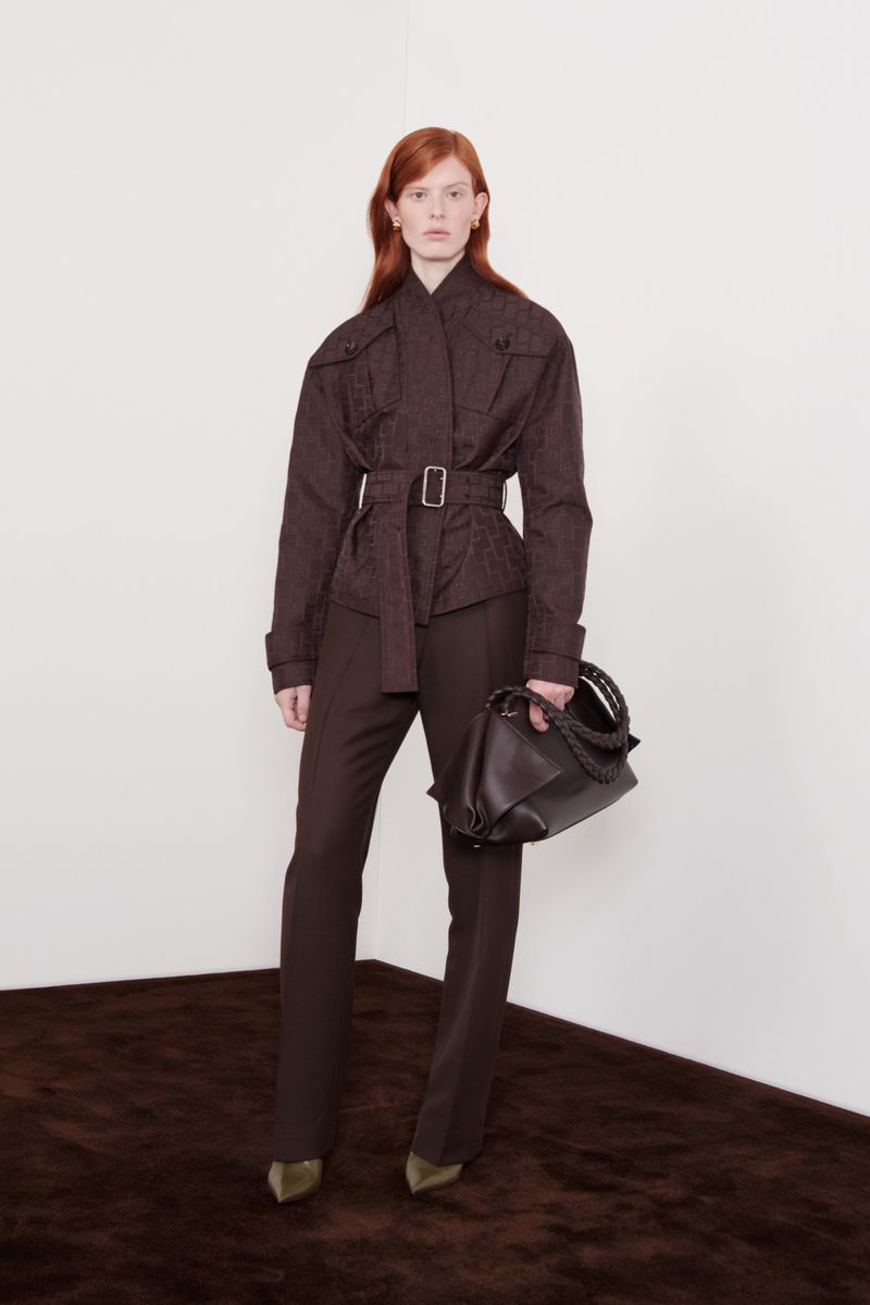A redhead woman poses indoors wearing a brown jacket, matching brown pants, and carrying a brown purse in a fashion editorial style image.