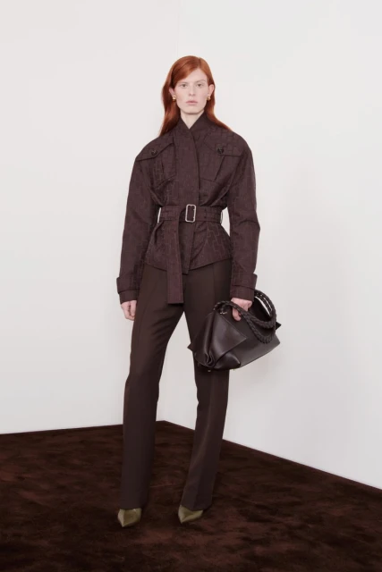 A redhead woman poses indoors wearing a brown jacket, matching brown pants, and carrying a brown purse in a fashion editorial style image.
