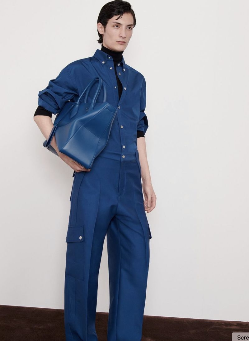 A person in a blue jumpsuit and a black turtleneck poses indoors while holding a blue leather tote bag against a white background.