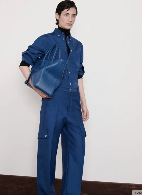 A person in a blue jumpsuit and a black turtleneck poses indoors while holding a blue leather tote bag against a white background.