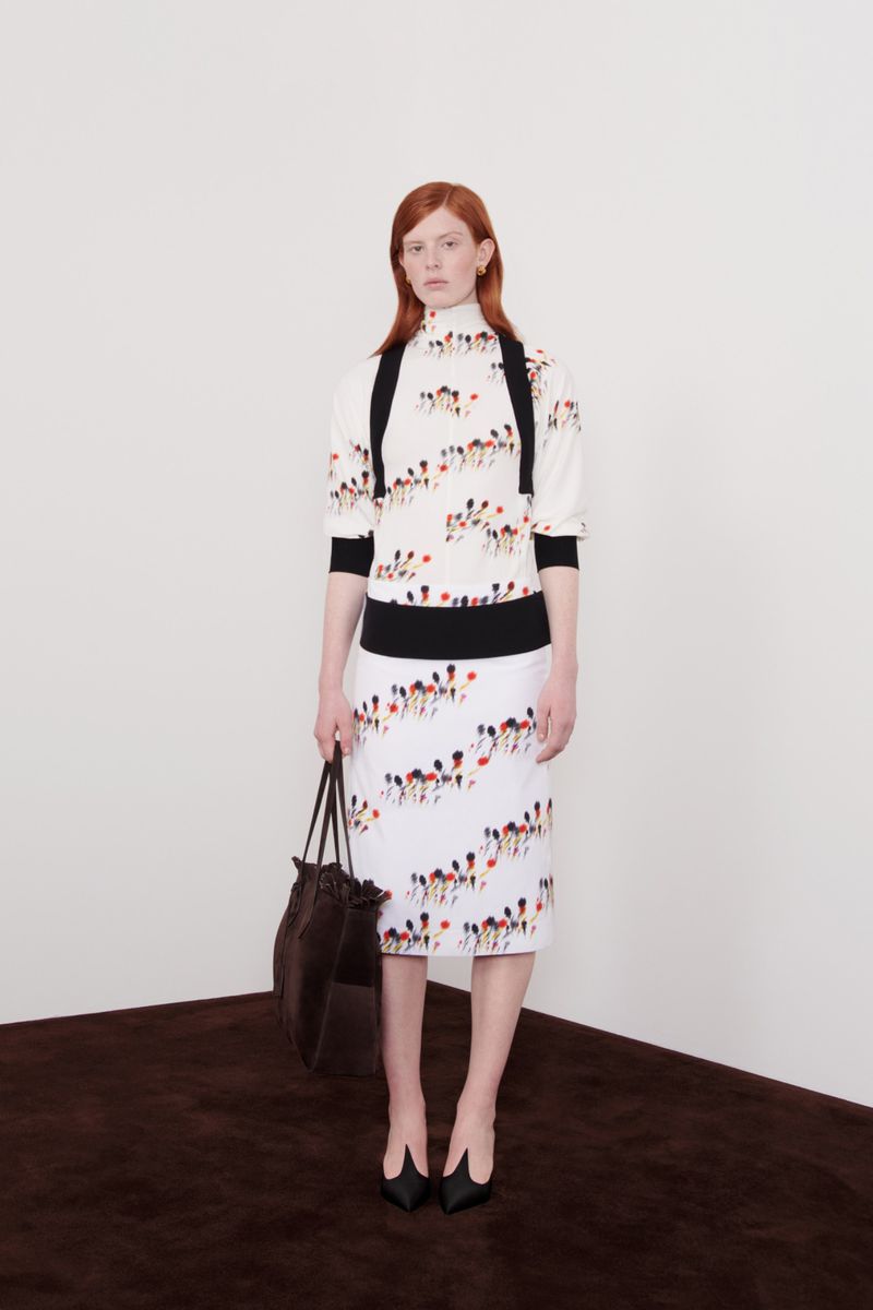 Fashion model posing in a studio wearing a white printed sweater and skirt with black heels and a large tote bag. Studio shot.