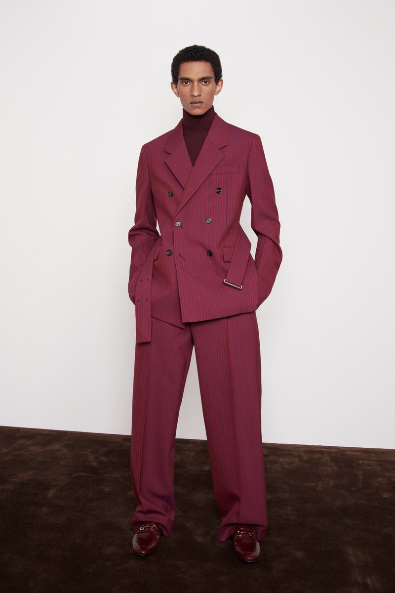 Fashionable man poses in a double-breasted maroon pinstripe suit with matching turtle neck and brown leather loafers against a white backdrop.
