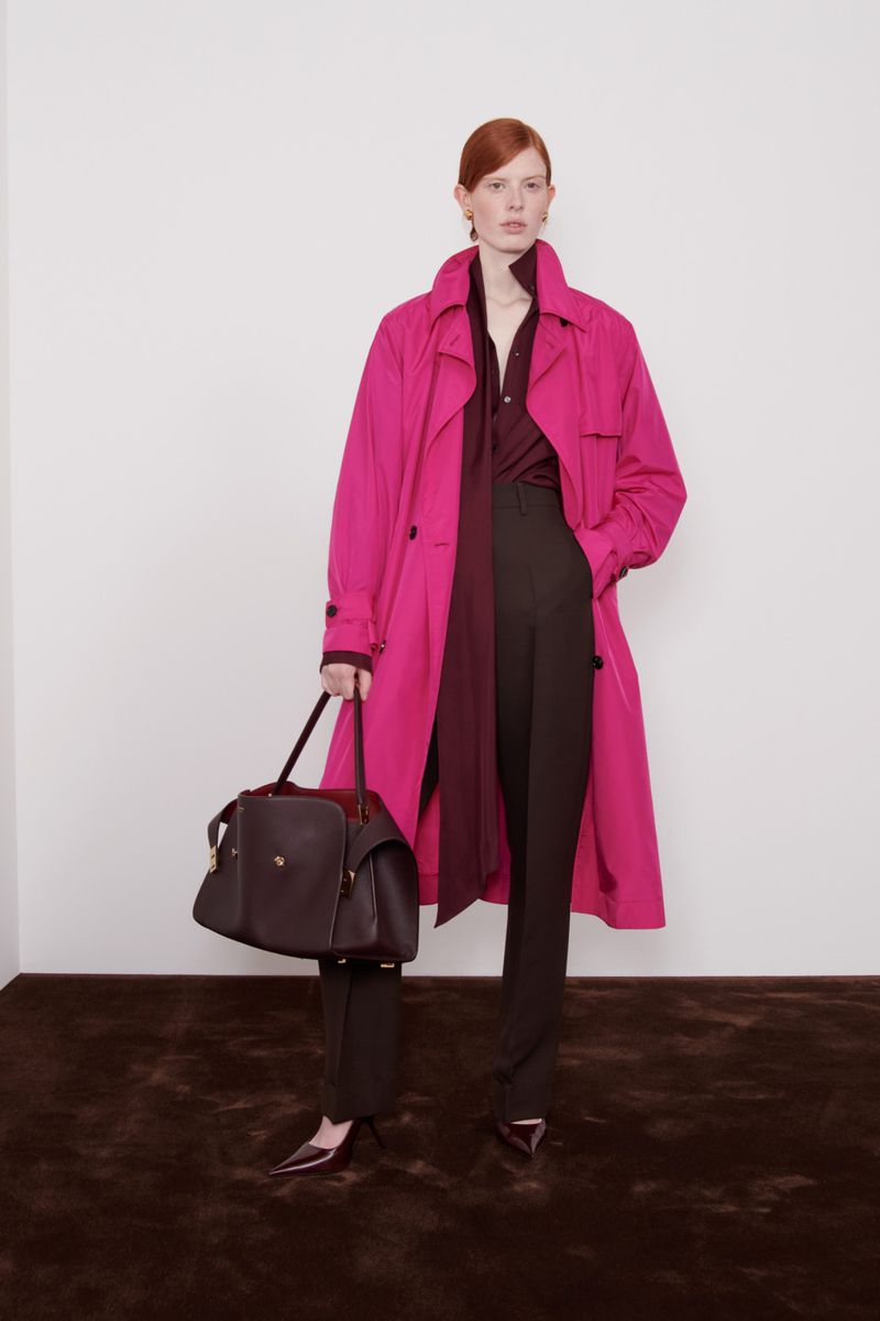 Fashionable woman in a pink trench coat and brown pants holding a dark brown leather handbag against a neutral backdrop.