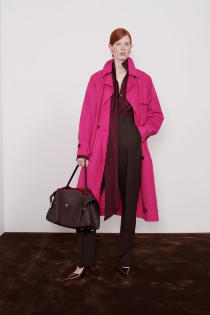 Fashionable woman in a pink trench coat and brown pants holding a dark brown leather handbag against a neutral backdrop.