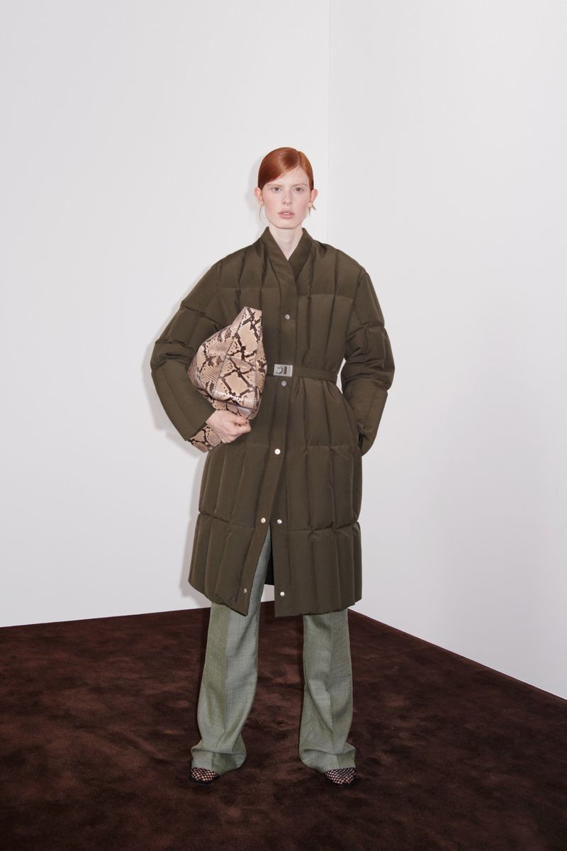 A redhead woman in a long, olive green quilted coat with a belt stands in front of a white wall, holding a snakeskin-patterned clutch.