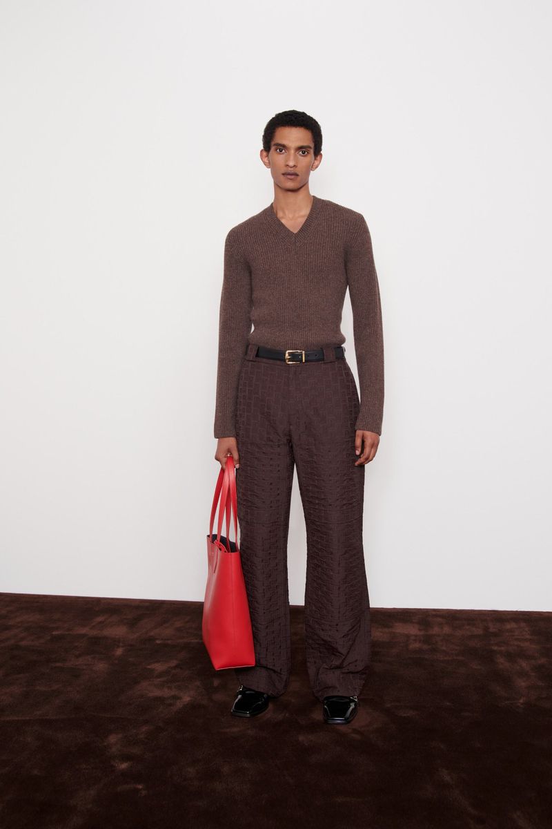 Man poses in brown sweater and trousers with red tote bag, standing on a dark rug against a white wall. Fashion editorial.