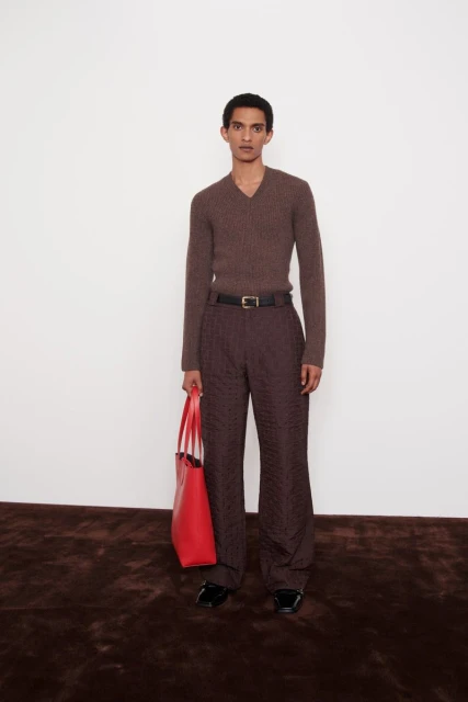 Man poses in brown sweater and trousers with red tote bag, standing on a dark rug against a white wall. Fashion editorial.