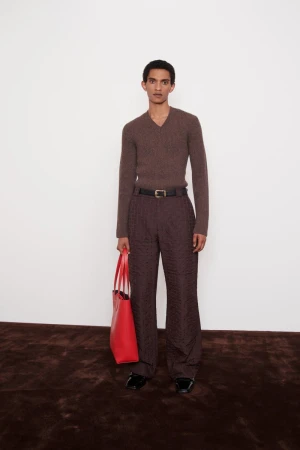 Man poses in brown sweater and trousers with red tote bag, standing on a dark rug against a white wall. Fashion editorial.