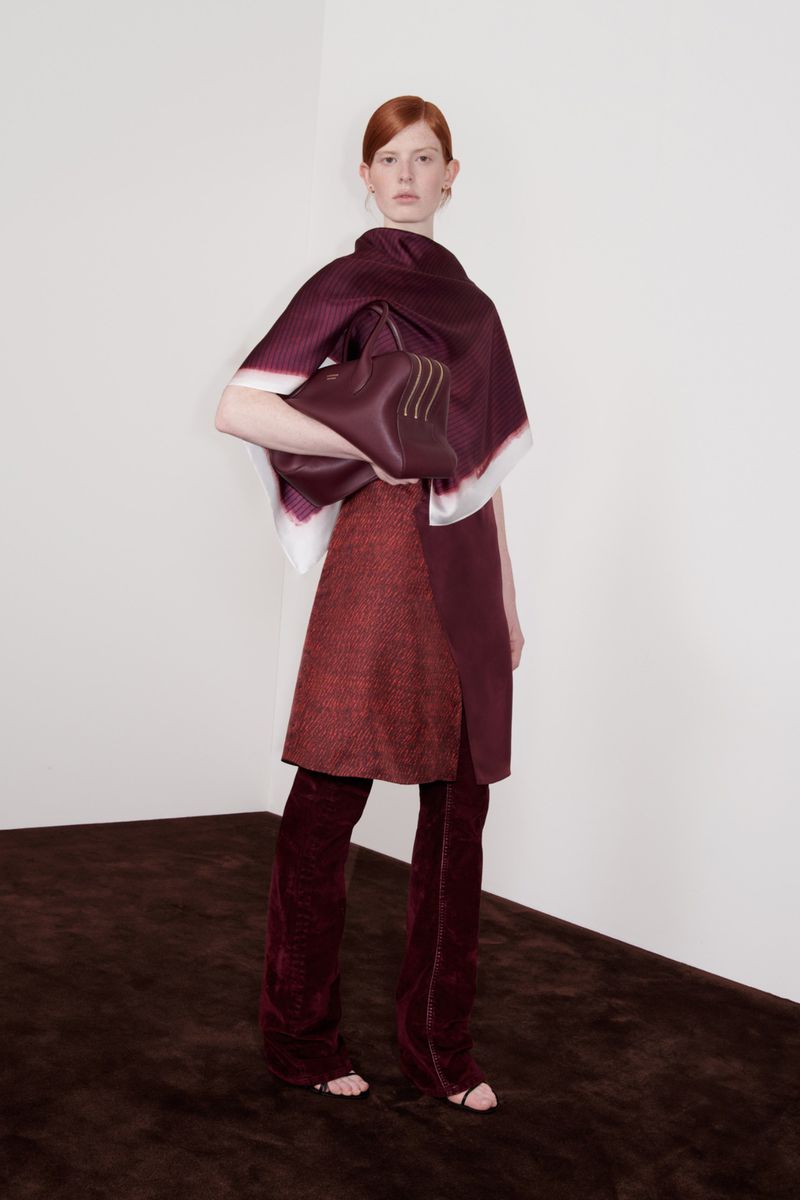 Fashion shot of a red-haired woman in a burgundy outfit holding a matching handbag, posing in a corner of a photo studio with a brown floor.