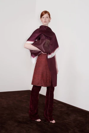 Fashion shot of a red-haired woman in a burgundy outfit holding a matching handbag, posing in a corner of a photo studio with a brown floor.