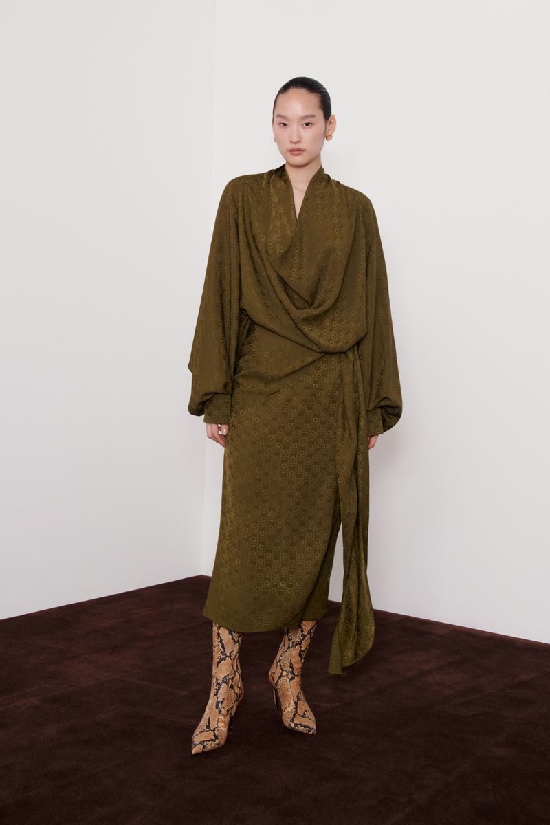 A stylish woman in an olive-green, patterned dress with long sleeves and snakeskin boots stands indoors against a white wall.