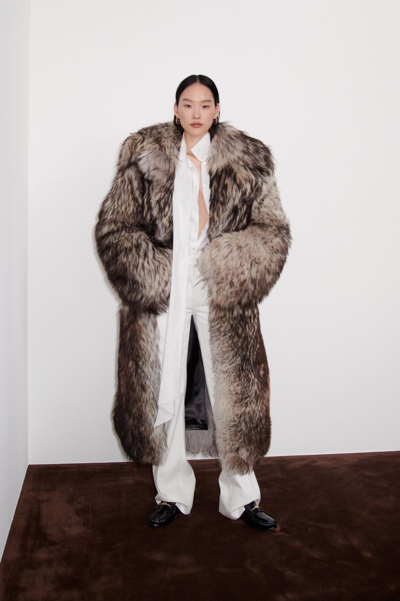 A slender Asian woman is captured in a full shot, wearing a long, striking fur coat, white pants and top, set against a white background.