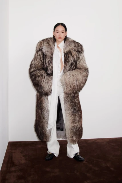 A slender Asian woman is captured in a full shot, wearing a long, striking fur coat, white pants and top, set against a white background.
