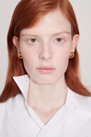 Close-up portrait of a woman with red hair wearing textured gold earrings and a white collared shirt against a white backdrop.