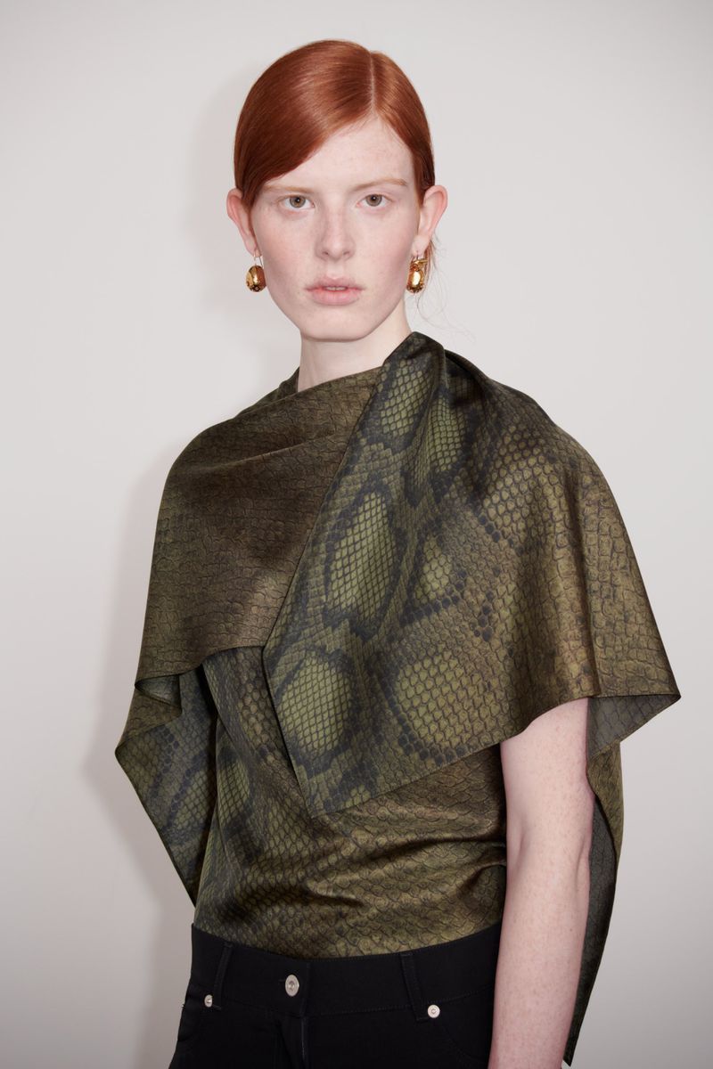 A red-haired woman in a snakeskin-patterned top poses in front of a neutral background, highlighting a fashion-forward and sophisticated look.