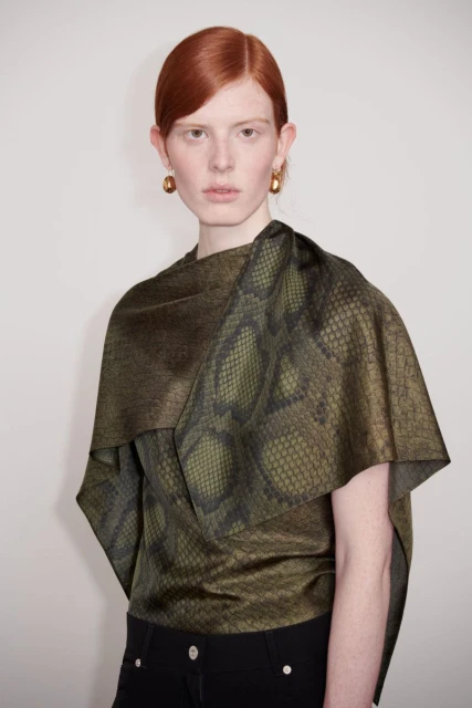 A red-haired woman in a snakeskin-patterned top poses in front of a neutral background, highlighting a fashion-forward and sophisticated look.