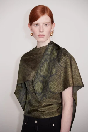 A red-haired woman in a snakeskin-patterned top poses in front of a neutral background, highlighting a fashion-forward and sophisticated look.