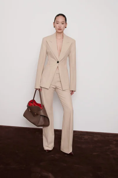 Model Yoonmi Sun wearing a tailored beige trouser suit and holding a brown leather handbag against a studio backdrop for Ferragamo.