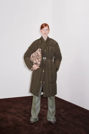 Model with red hair posing in an olive green quilted belted coat, light green trousers, and a snakeskin clutch bag.