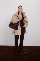 Model wearing a beige jacket and dark trousers holding a black leather shoulder bag against a plain studio background.