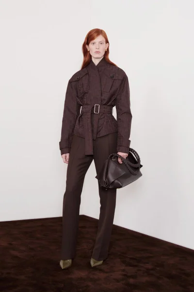 Model Laura Rudd wears a dark brown belted jacket and tailored trousers while holding a leather handbag for Ferragamo Resort 2025.
