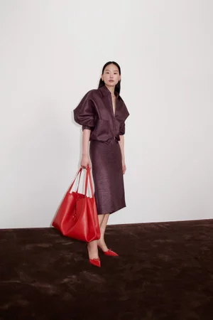 Model in plum textured matching jacket and skirt holding a large red tote bag against a plain wall and dark floor